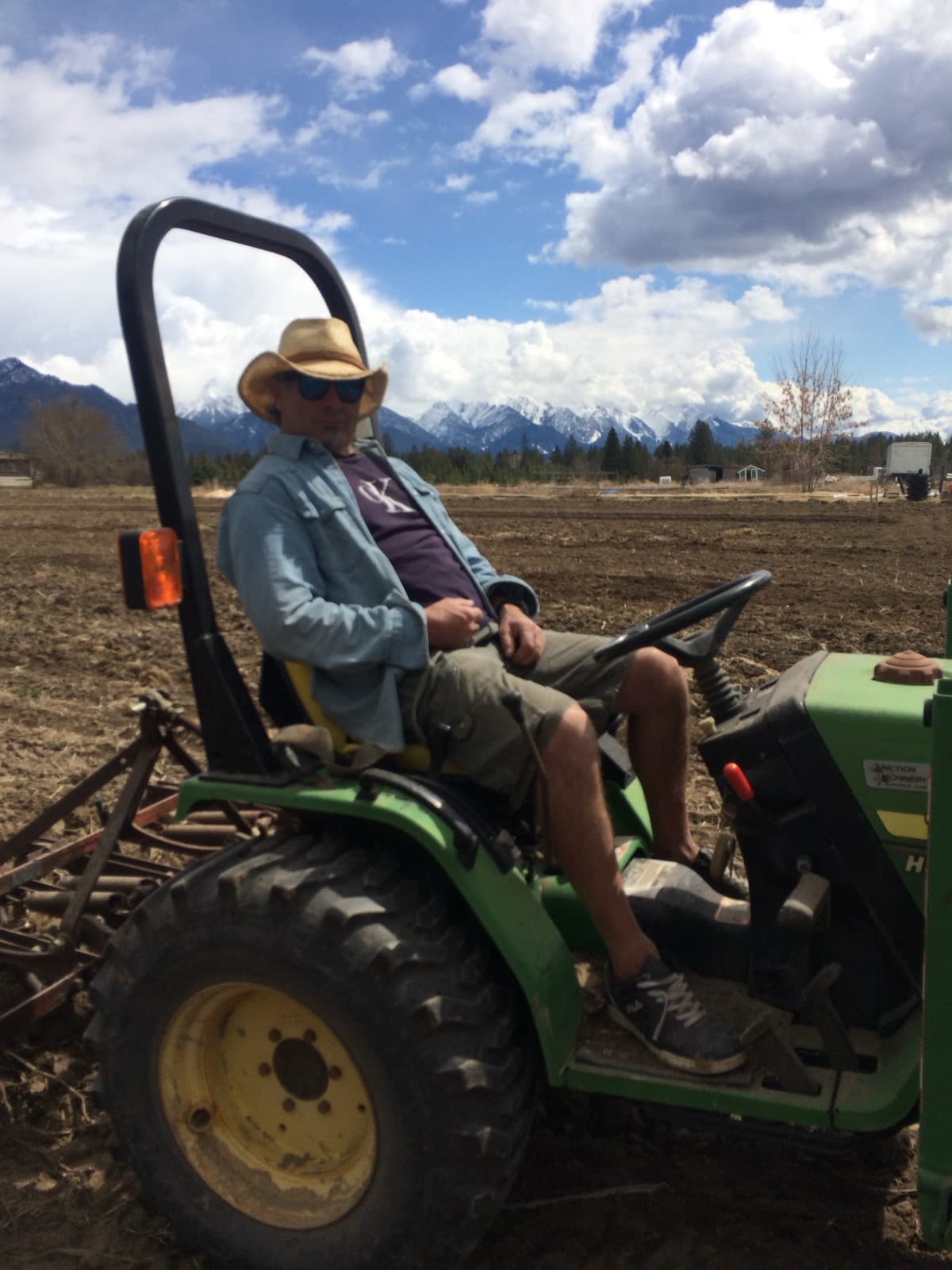 Peter, Tanner's dad, on a green compact tractor in a freshly tilled field, straw hat and sunglasses, snow-capped mountains behind him